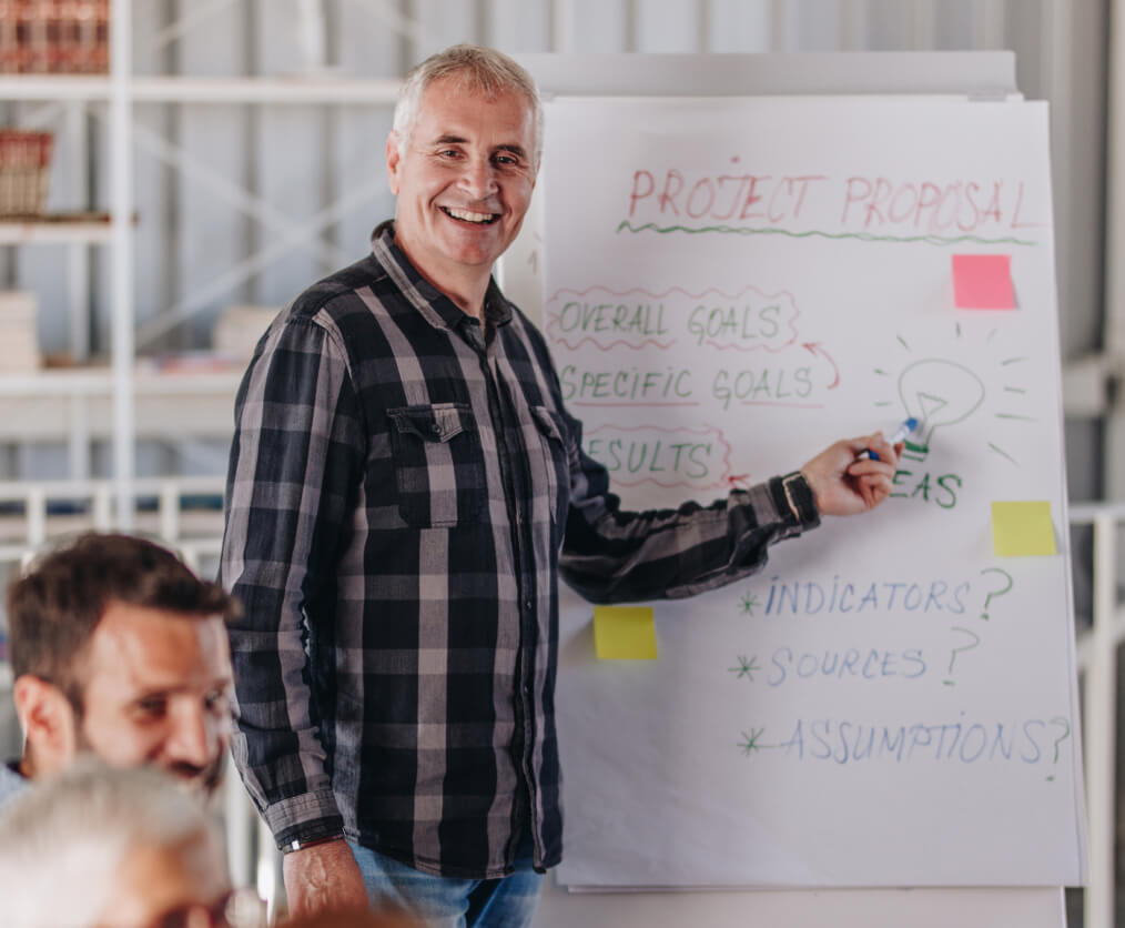 A man in a plaid shirt smiling and pointing project proposal notes in front of a table of people