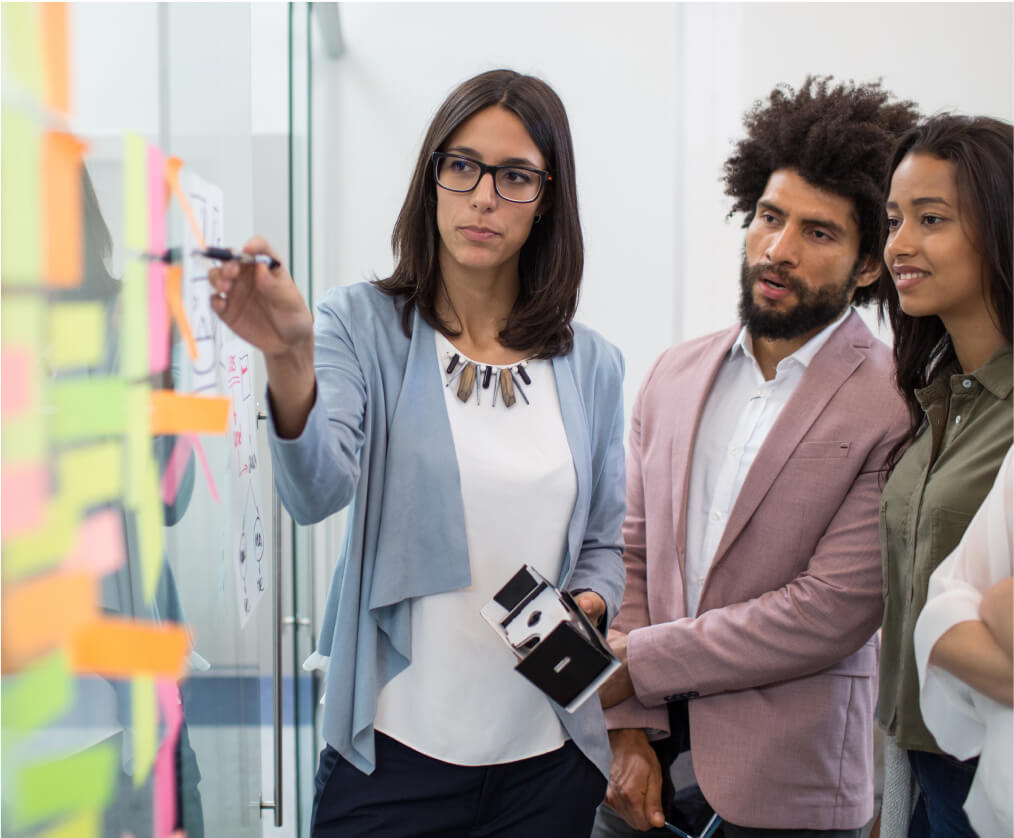 A group of three people standing and looking at a white board covered in colored sticky notes while one woman points to one with a pen