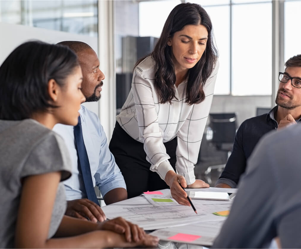 A group of people sitting around a table while a woman stands pointing down to paperwork on the table